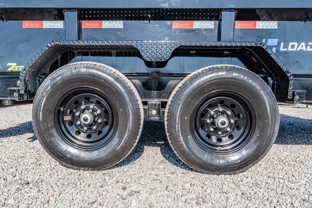 Dual trailer wheels with black rims on a dark blue dump trailer, parked on gravel ground
