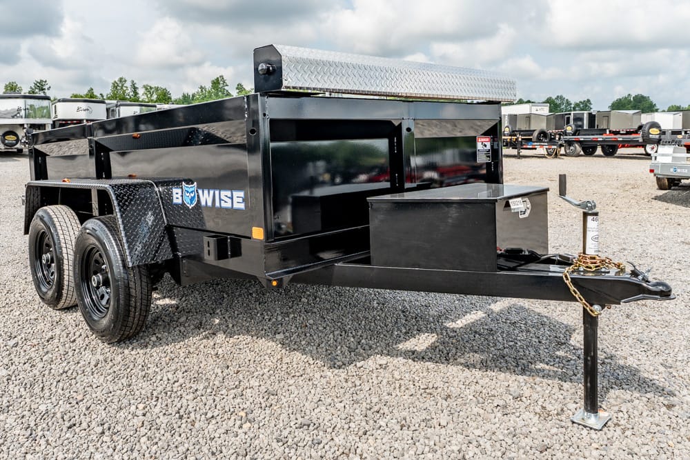 Black dual-axle dump trailer on a gravel lot, featuring a diamond-plate toolbox and front hitch with safety chain (tow-ready).