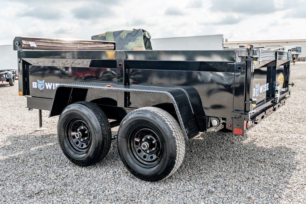 Black tandem-axle dump trailer on a gravel lot, with tall metal sides and a tarp cover on top.