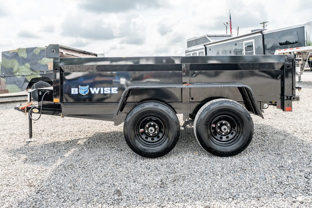 Black dual-axle dump trailer with the B-WISE logo parked on a gravel lot; camouflage-covered equipment and other trailers visible in the background.