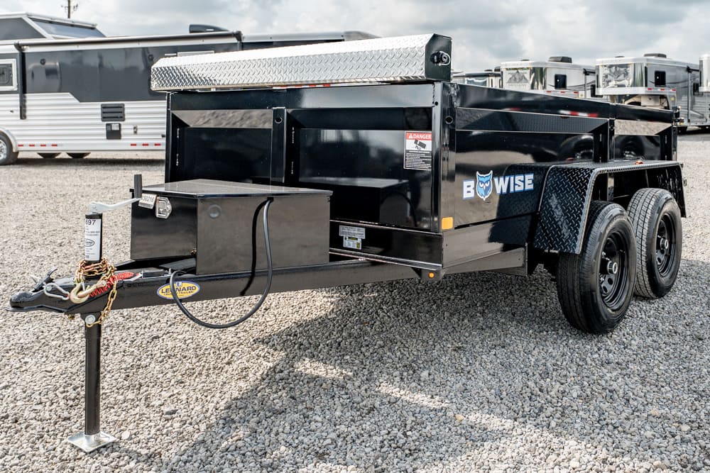 Black Bow-Wise dual-axle utility trailer on a gravel lot, with a front toolbox, hitch jack, and diamond-plate accents; RVs in the background