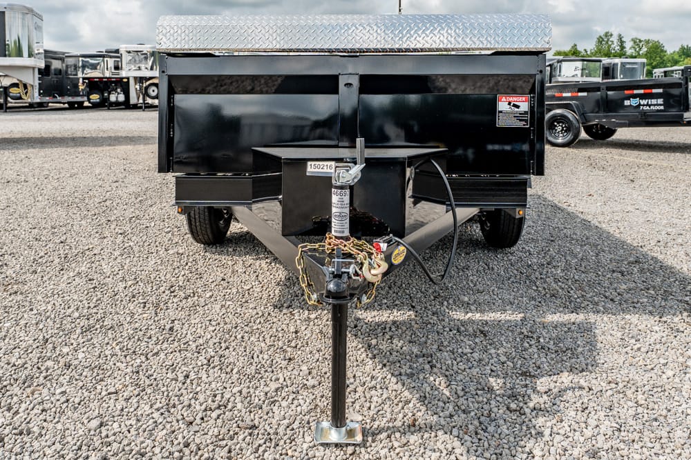 Front view of a black utility dump trailer on a gravel lot, connected to a hitch with safety chains and the jack stand extended; diamond-plate lid visible on top.