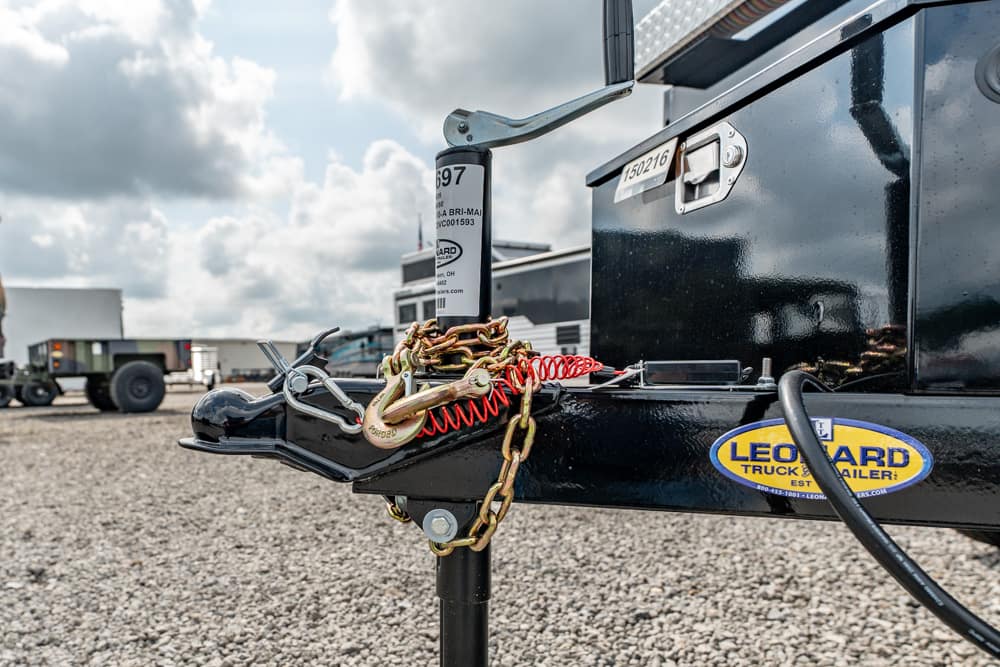 Close-up of a trailer hitch with safety chains and a red coil spring on a black trailer, in a truck yard.