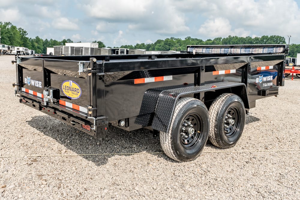 Black two-axle dump trailer on a gravel lot with orange-white reflectors and diamond-plate fenders, showing brand stickers on the sides.