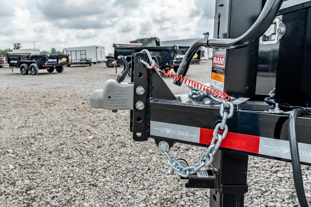Close-up of a trailer hitch with a padlock-style coupler lock, safety chains, and a red coiled cable on a gravel lot.