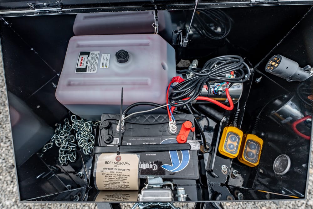 A battery and wiring setup inside a black storage compartment, with a translucent fuel jug, chains, and red and black cables coiled nearby.