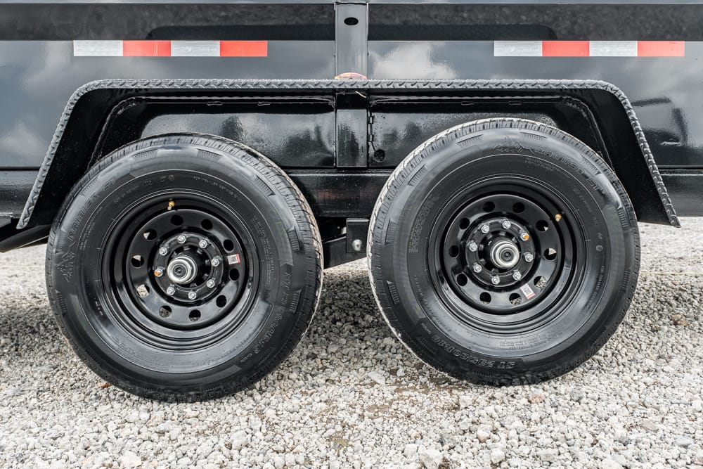 Dual-axle trailer with two black steel wheels and tires under a black trailer body on a gravel surface.