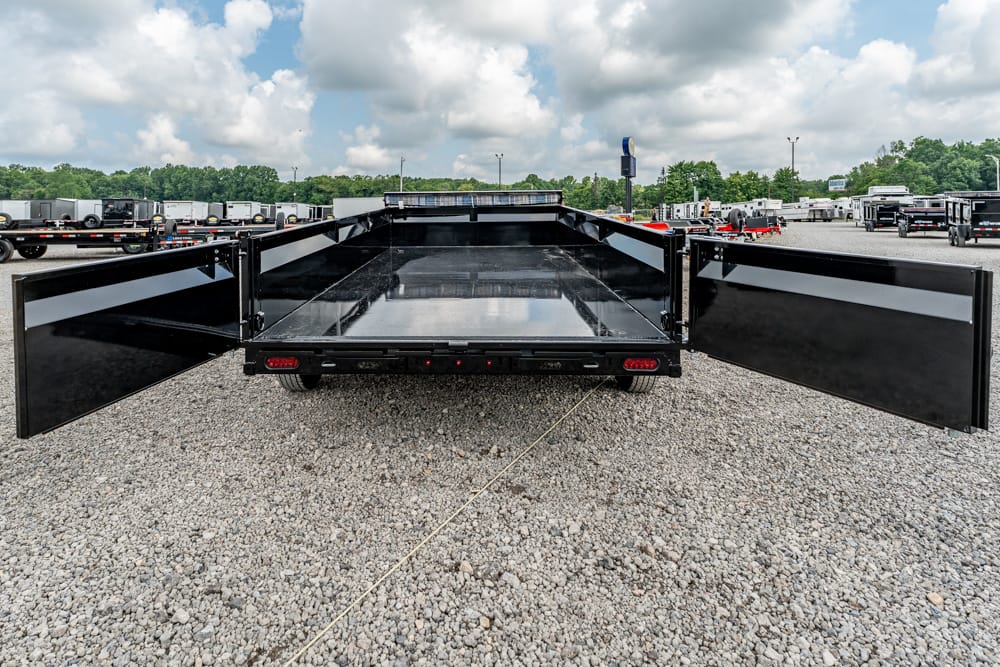 Black dump trailer with its dual rear doors wide open, showing an empty cargo bed on a gravel lot among many trailers and trucks under a cloudy sky