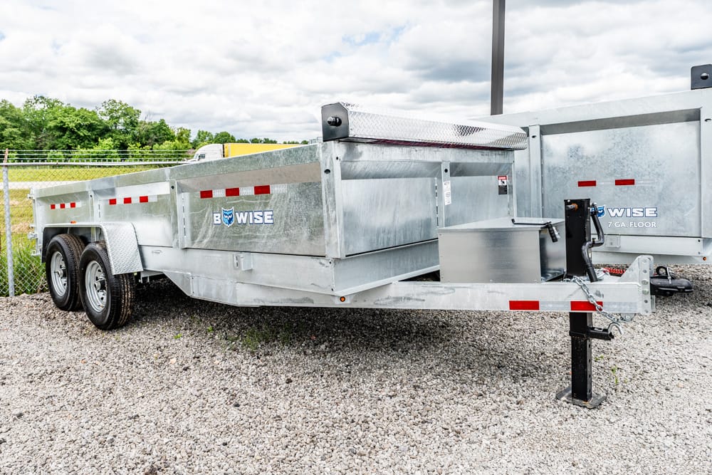 Dump trailer on a gravel lot with dual axles and a front hitch, branded BowWise. Used for hauling materials on a construction site.