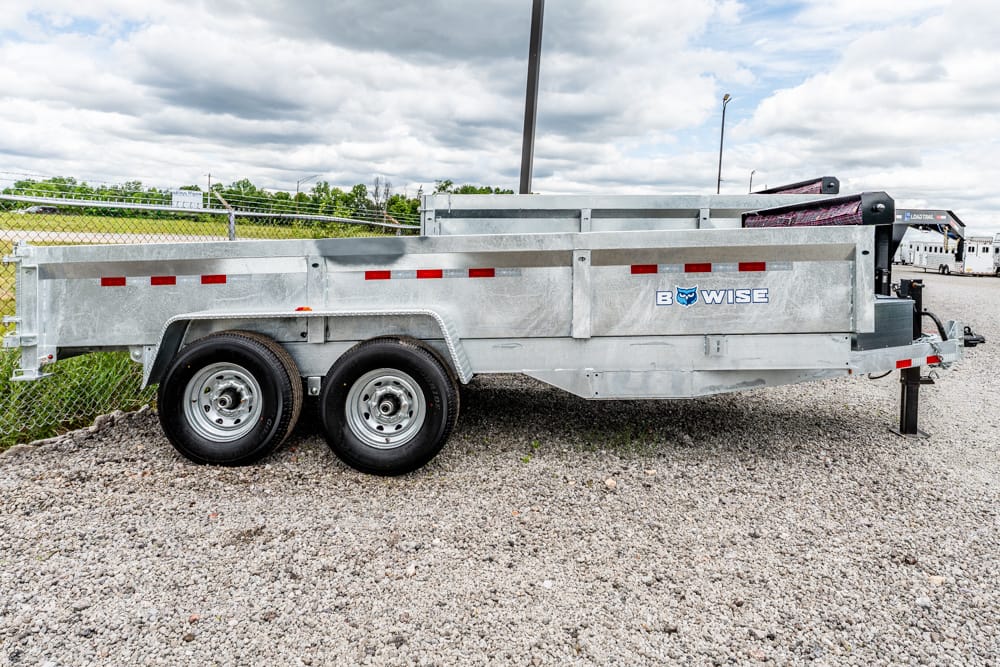 Side view of a silver B-WISE dump trailer with dual rear tires on a gravel lot, ready for use.