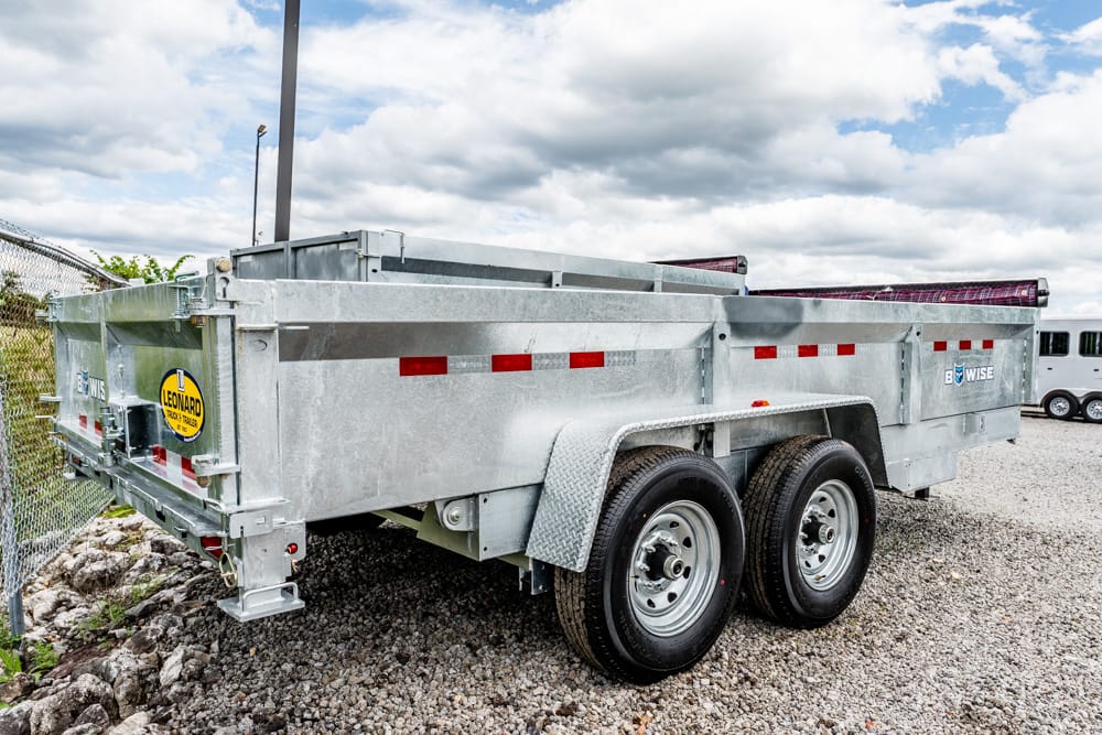 Twin-axle metal dump trailer with high side walls on a gravel lot, showing red reflective strips and a Leonard logo on the back end.