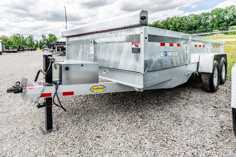 Silver tandem-axle utility trailer on a gravel lot, hitched to a truck with safety chains and a jack stand, branded B-WISE.
