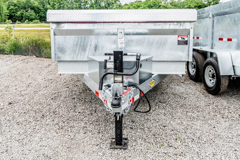 Front view of a metal utility trailer on a gravel lot, showing the hitch, safety chains, and jack stand.