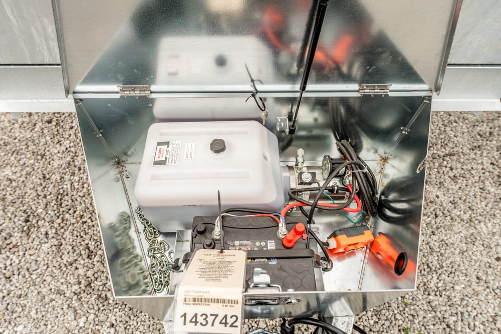Open metal housing containing a white plastic fuel tank, a battery with red/black leads, and orange clamps on a gravel surface (electrical equipment setup).