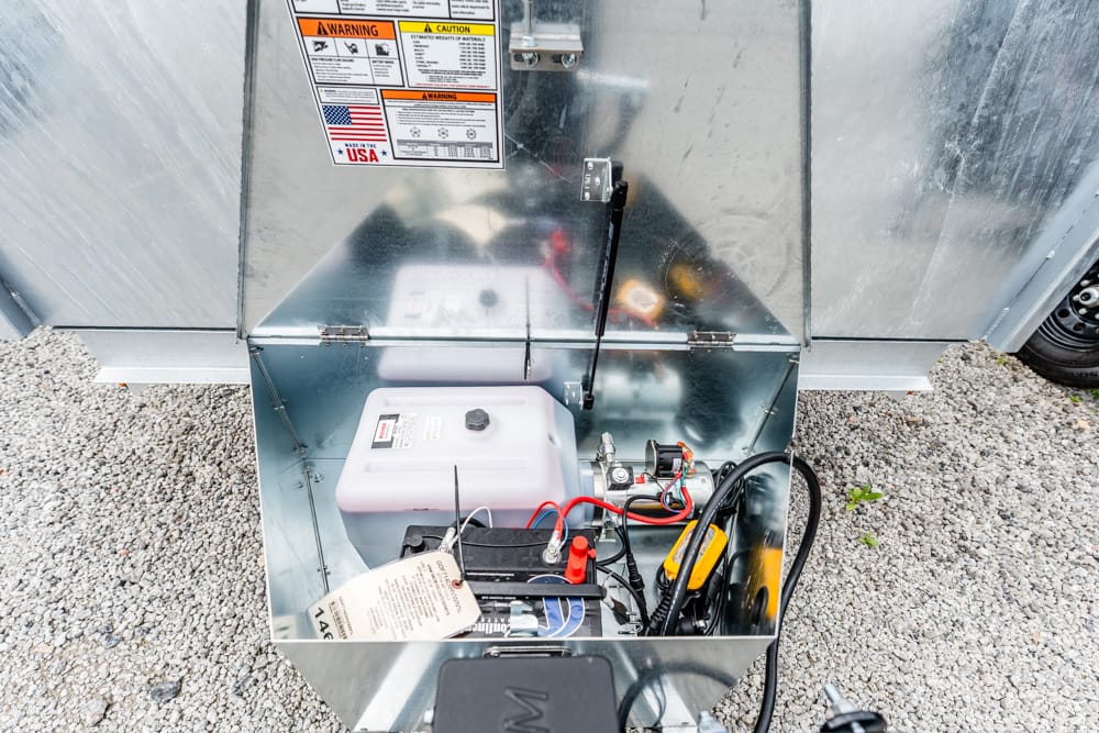 Open metal cabinet housing a small generator; visible components include a white fuel container, red/black battery cables, and warning labels on the lid.