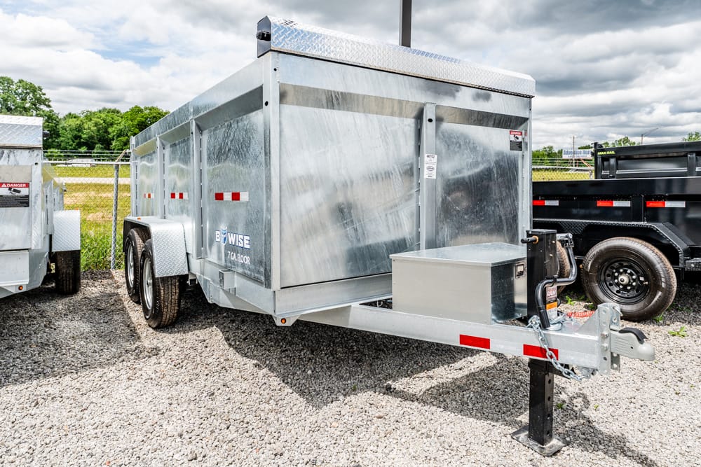 Two‑axle silver metal cargo trailer with a front storage box, parked on a gravel lot.