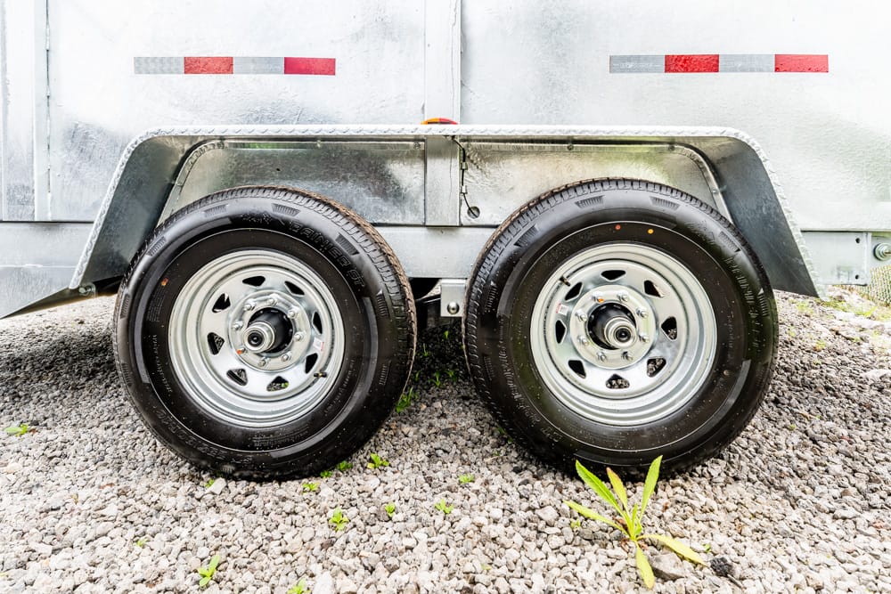 Dual-axle trailer wheels with silver rims on a gravel surface under metal fenders.