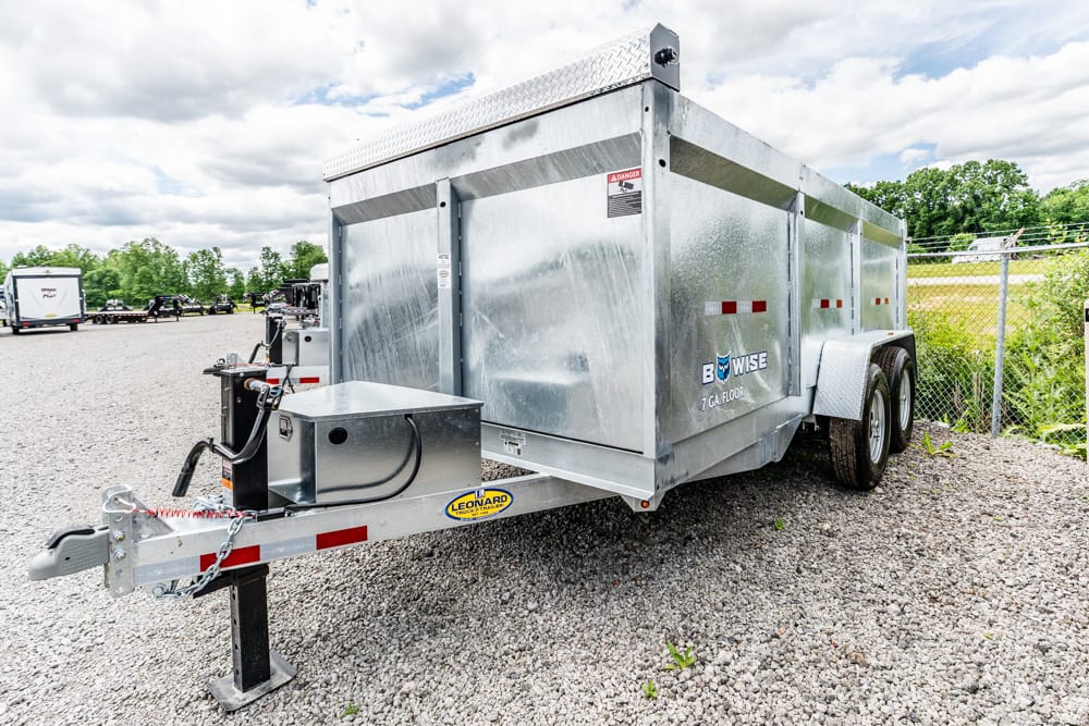 Silver aluminum enclosed dump trailer on a gravel lot, front hitch and safety chains visible, dual rear wheels, fence in background.
