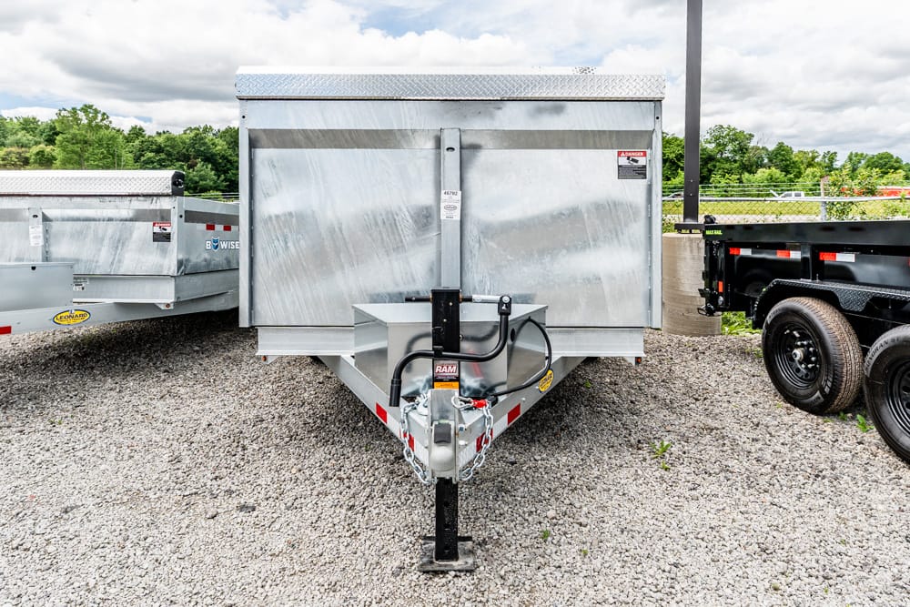 Front view of a silver metal utility trailer on a gravel lot, showing the hitch, jack, and safety chains.