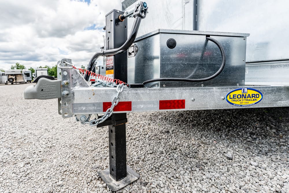 Close-up of the front of a trailer showing a stabilizing jack, hitch, safety chains, and a Leonard Truck & Trailer decal on a gravel lot.