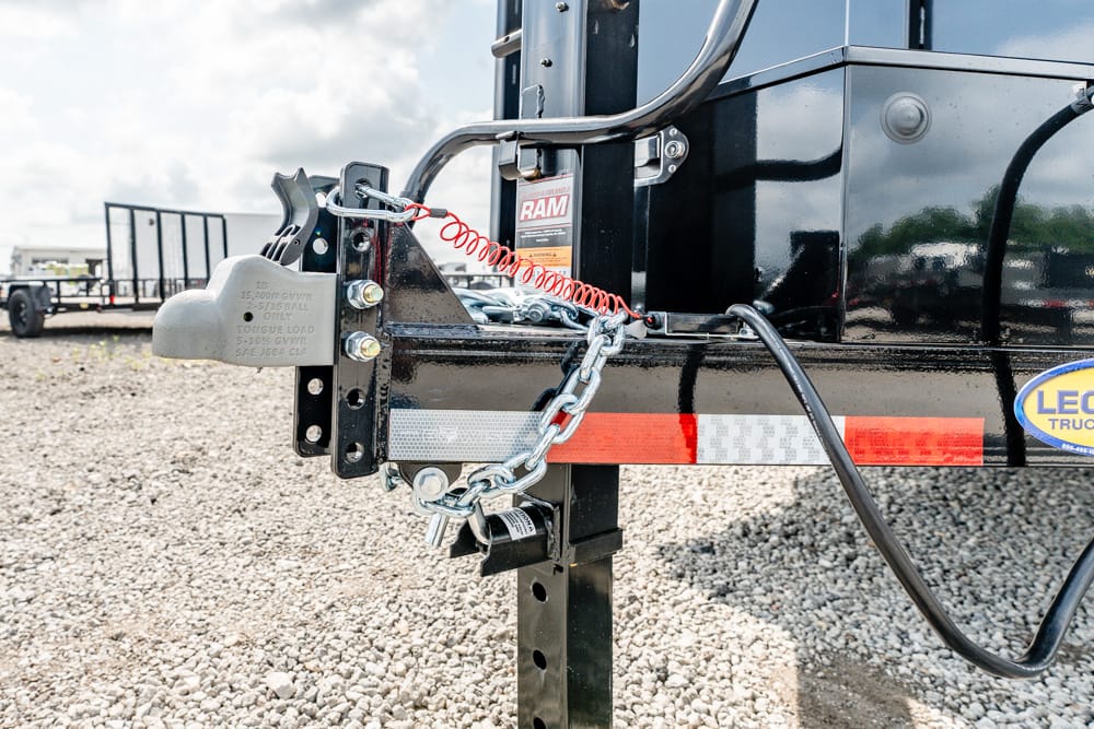 Close-up of a trailer hitch latch, safety chain, and red coiled lock securing a black utility trailer on a gravel lot.