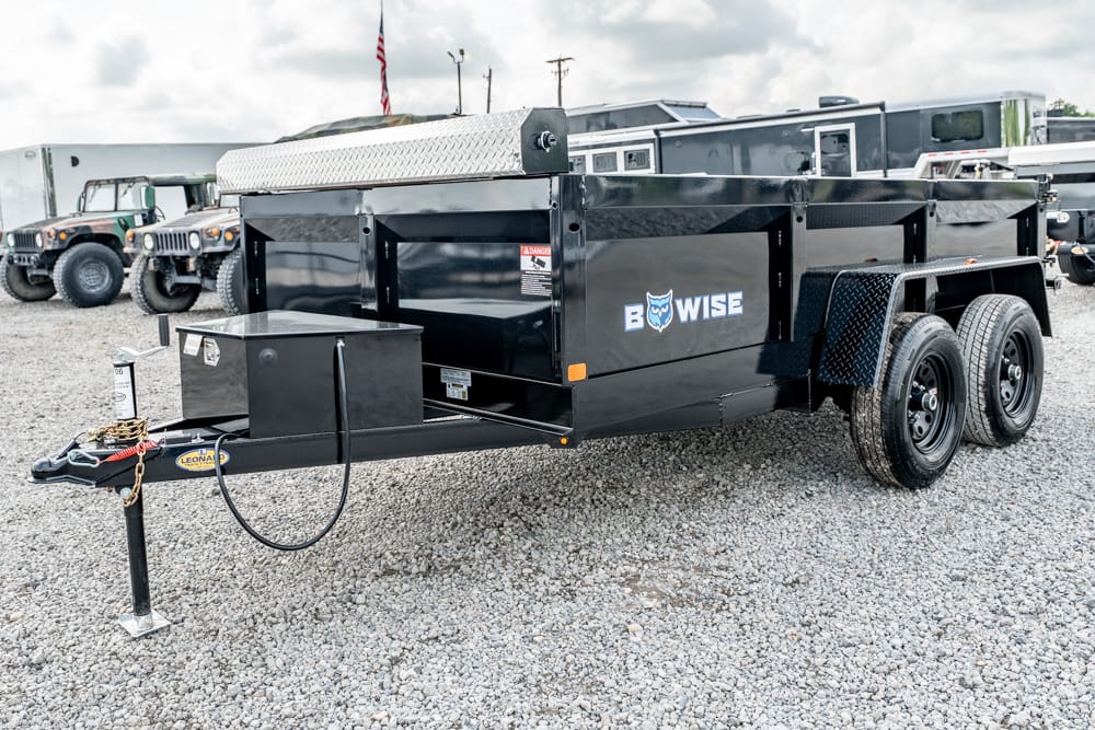 Black tandem-axle dump trailer on a gravel lot with a front hitch, safety chains, and a toolbox area visible. (Informative)