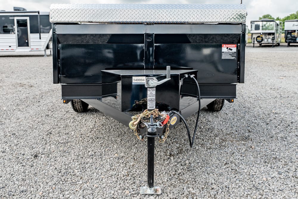 Black utility trailer hitch close-up with jack, safety chains, and wiring on a gravel lot; diamond-plate edge visible at top.