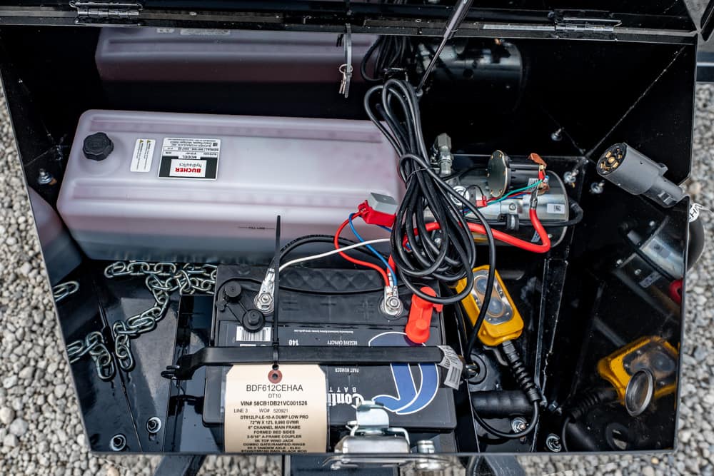 Inside a metal storage box: a car battery with red/black cables and assorted wires, a plastic battery box, and electrical components.