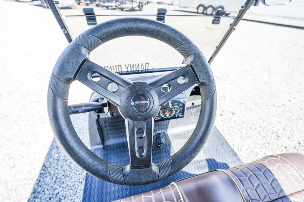 Steering wheel of a golf cart with visible dashboard gauges and brown leather seat nearby, outdoors on a gravel lot