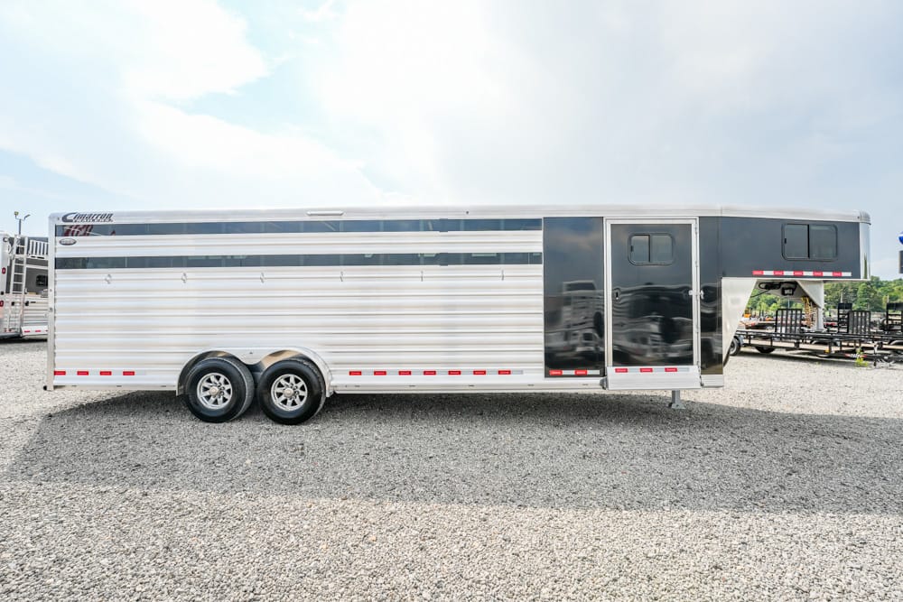 Long aluminum horse trailer with two axles parked on a gravel lot. The trailer has a silver body with a dark front section and a gooseneck area at the right.
