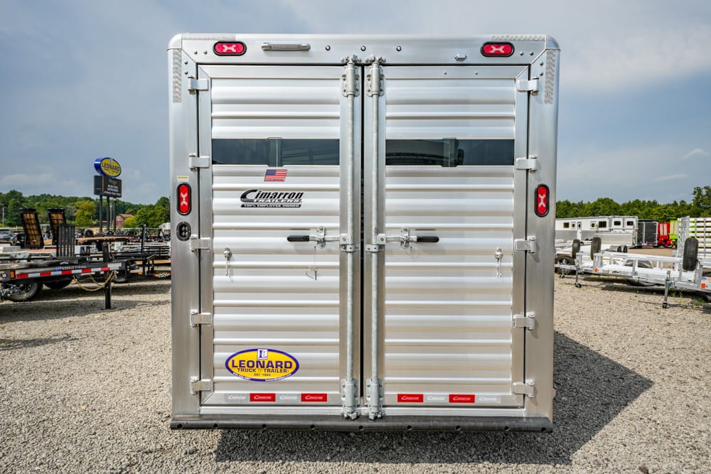 Rear view of a silver Cimarron trailer with double doors, latches, and red taillights on a gravel lot, featuring an American flag decal and Leonard Trailer logo.