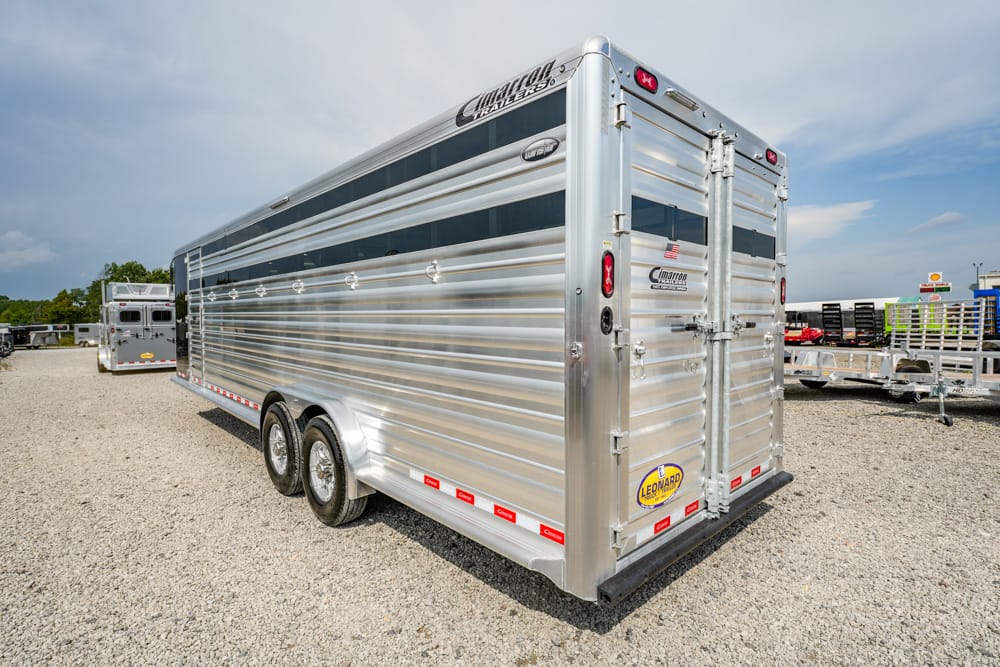 Silver aluminum trailer with two axles parked on a gravel lot; closed rear doors and cargo logos visible.