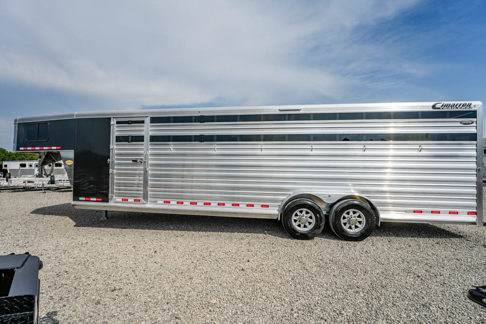 Silvery two-axle livestock trailer parked on a gravel lot under a blue sky with clouds, side doors closed and windows along the top edge