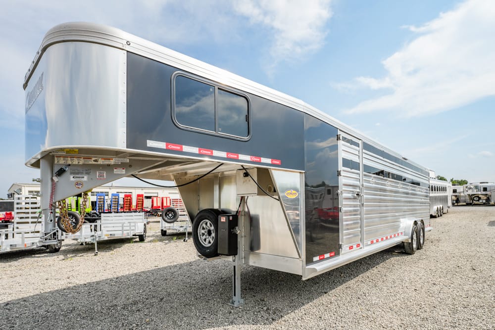 Silver gooseneck horse trailer with living quarters, parked on a gravel lot; side panels, windows, and a spare tire visible.