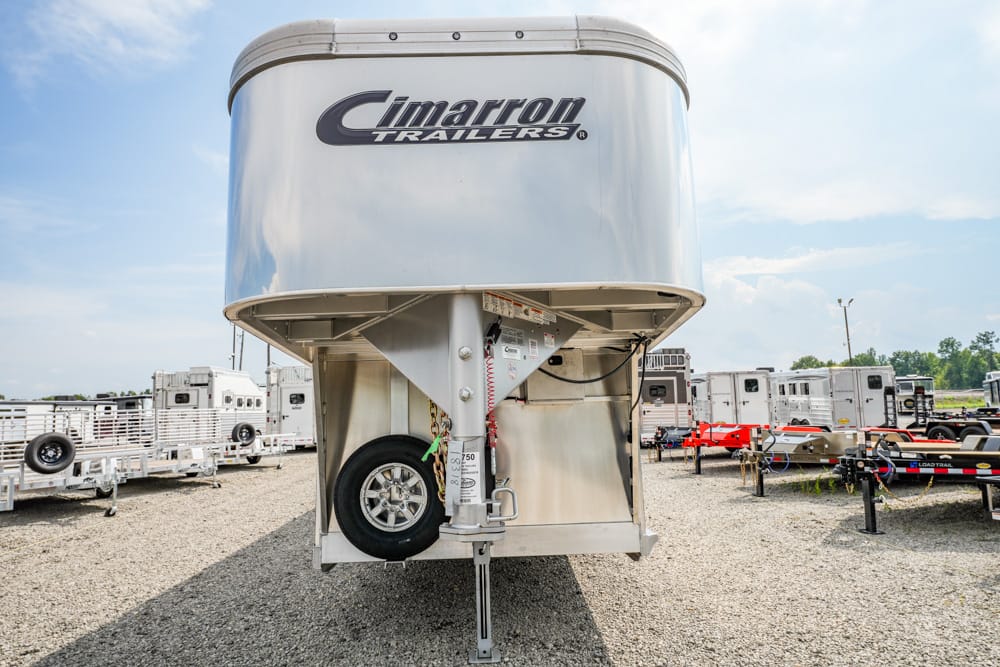 Silver Cimarron gooseneck trailer viewed from the front, showing the hitch, jack, spare tire, and 'Cimarron Trailers' logo, parked on gravel with other trailers in the background.