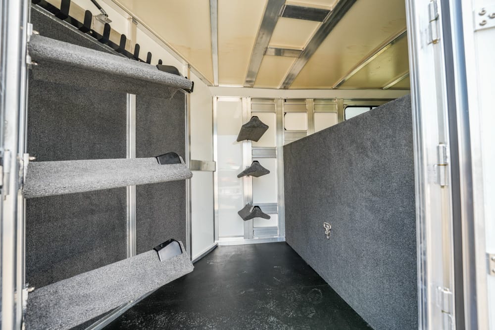 Interior of a horse trailer with wall-mounted saddle racks along the left and a padded divider on the right, and a dark floor in the center aisle.