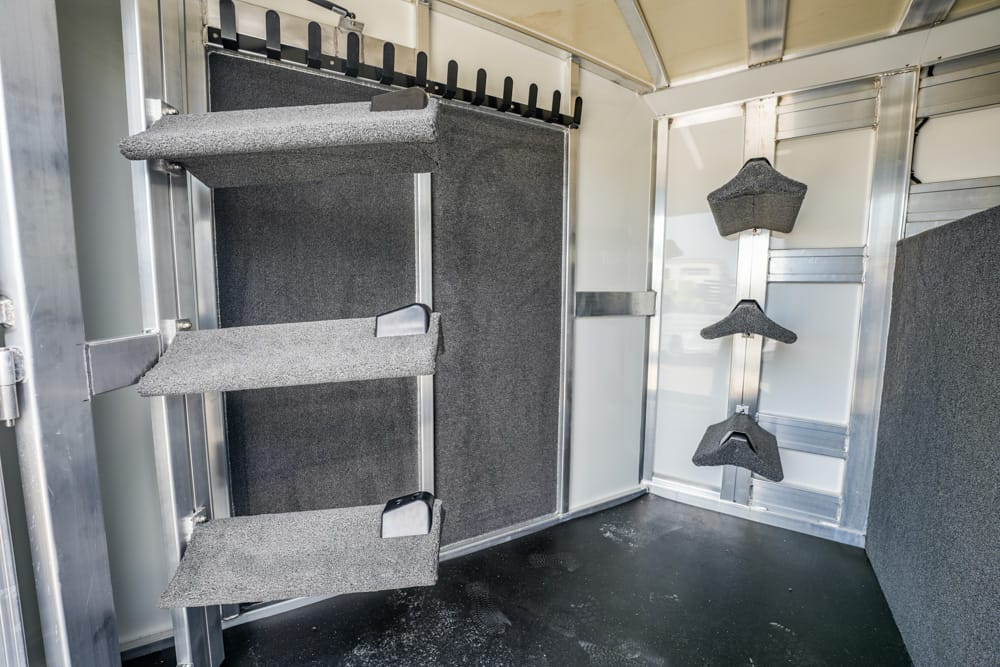 Interior of a horse trailer with left-side saddle racks and right-side padded tack hangers on metal walls.