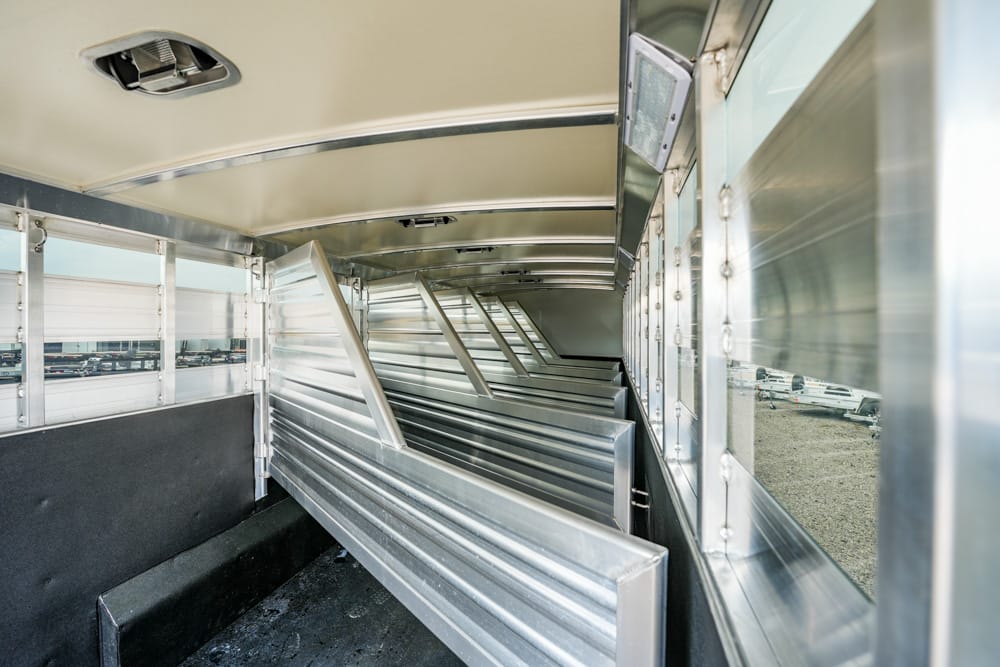 Interior of a shiny aluminum livestock trailer with partitioned stalls and windows along the side, parked vehicles visible outside.