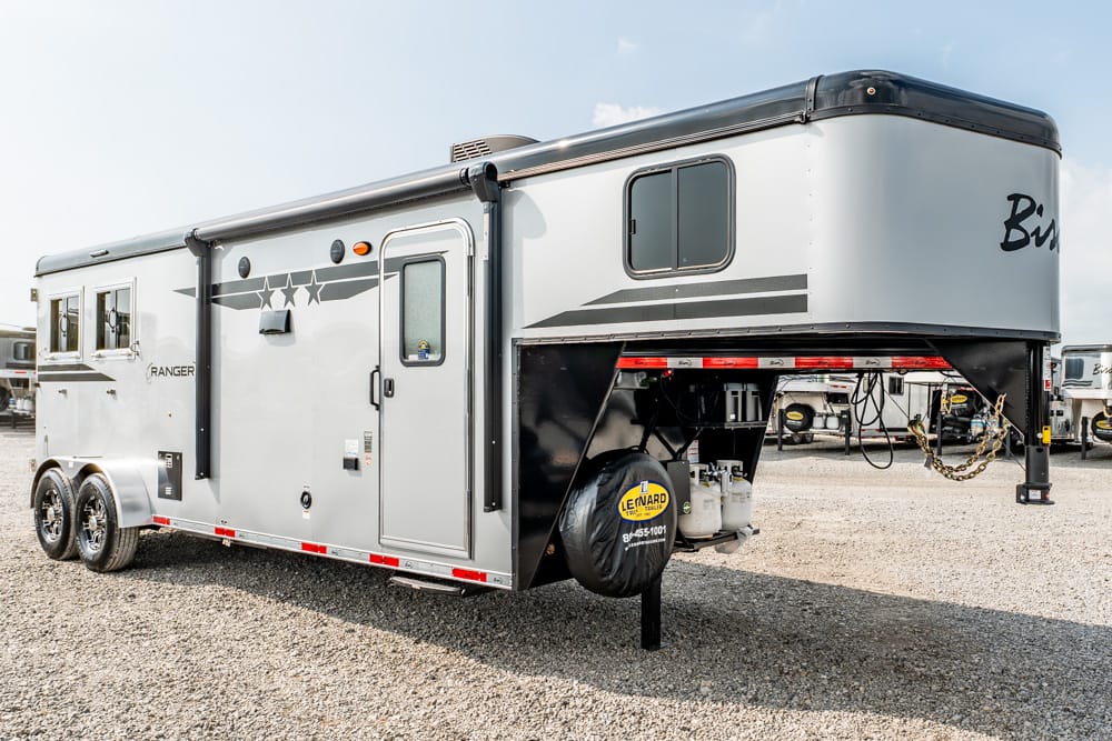 Silver triple-axle fifth-wheel RV/travel trailer parked on a gravel lot, front hitch raised and side door visible.