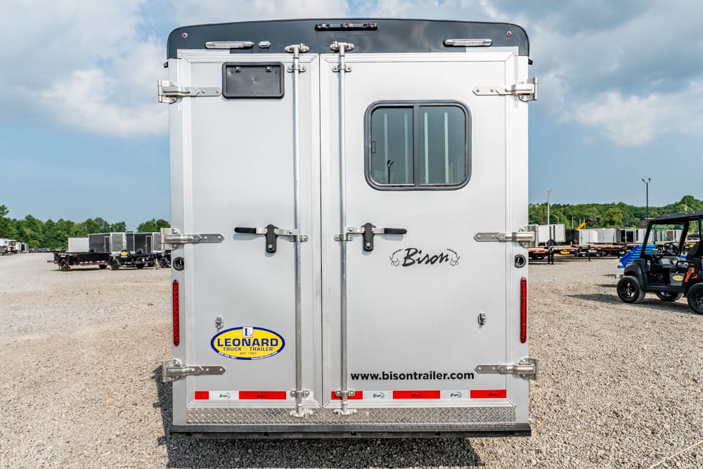 Rear view of a silver Bison livestock trailer with double doors, a small window on the right door, and branding stickers (Leonard Trailer) visible on a dealership lot.