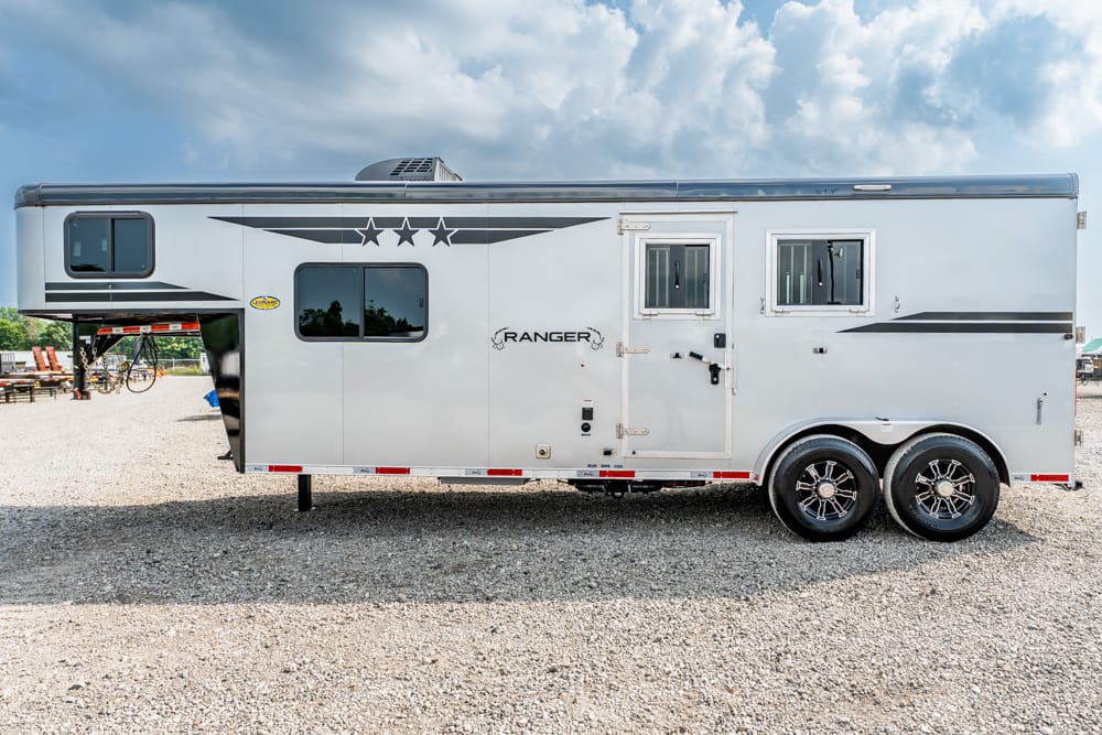 White gooseneck horse trailer with dual axles and black decorative stripes parked on a gravel lot beneath a cloudy sky, side view showing windows and entry door.