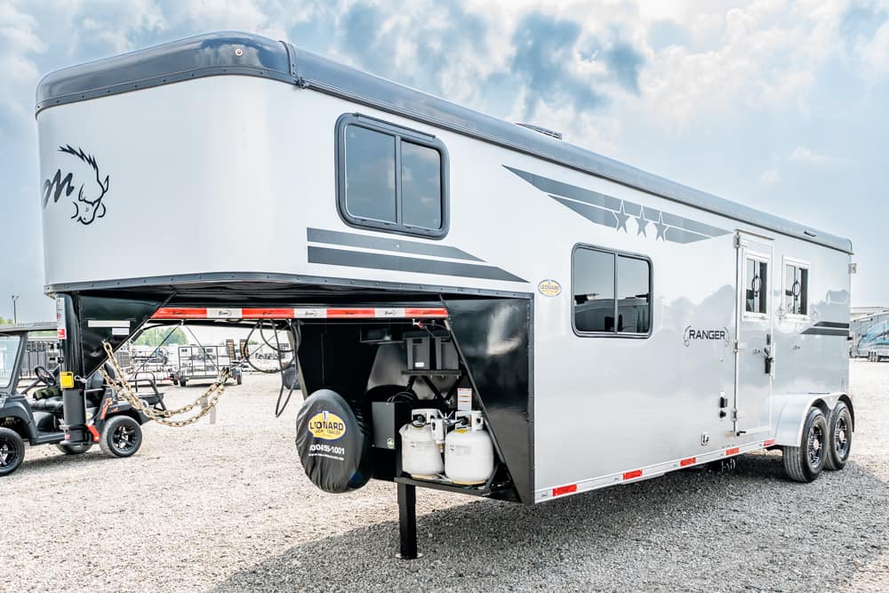 Silver Ranger travel trailer on a gravel lot, front storage area open with propane tanks and a spare tire visible.