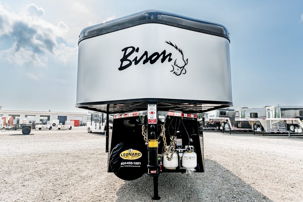 Front view of a large white Bison gooseneck trailer in a gravel lot, with spare tire and propane tanks under the hitch visible.