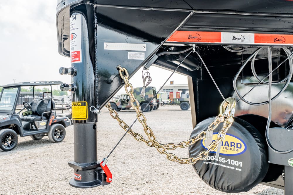 Close-up of a black trailer hitch with safety chains, red jack lever, and a spare tire cover on a gravel lot; background shows utility vehicles and equipment.