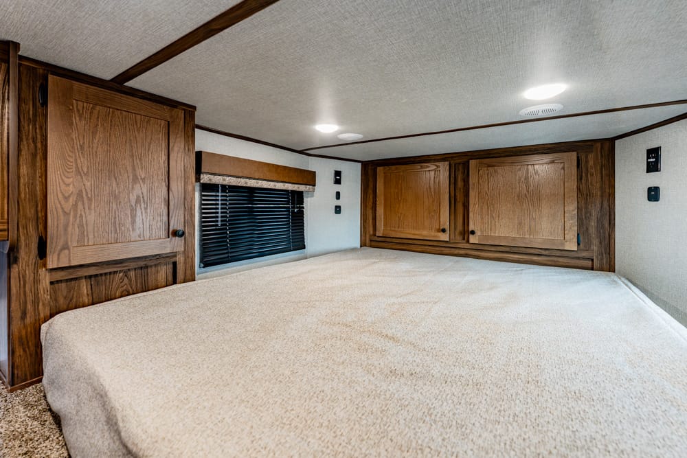 Wood-paneled RV bedroom with a large bed, overhead cabinets, and a window with blinds on the left.