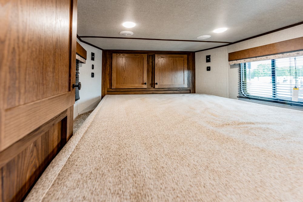 Bedroom inside a motorhome with a large beige bed, dark wood cabinets at the head, and a window with blinds on the side