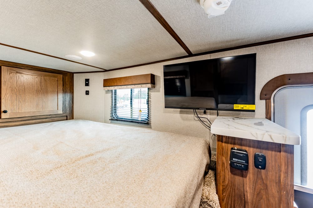 RV bedroom with a large bed, mounted flat-screen TV, and wood cabinetry along the wall. window with blind to the left. Laminated nightstand and outlets near the TV.
