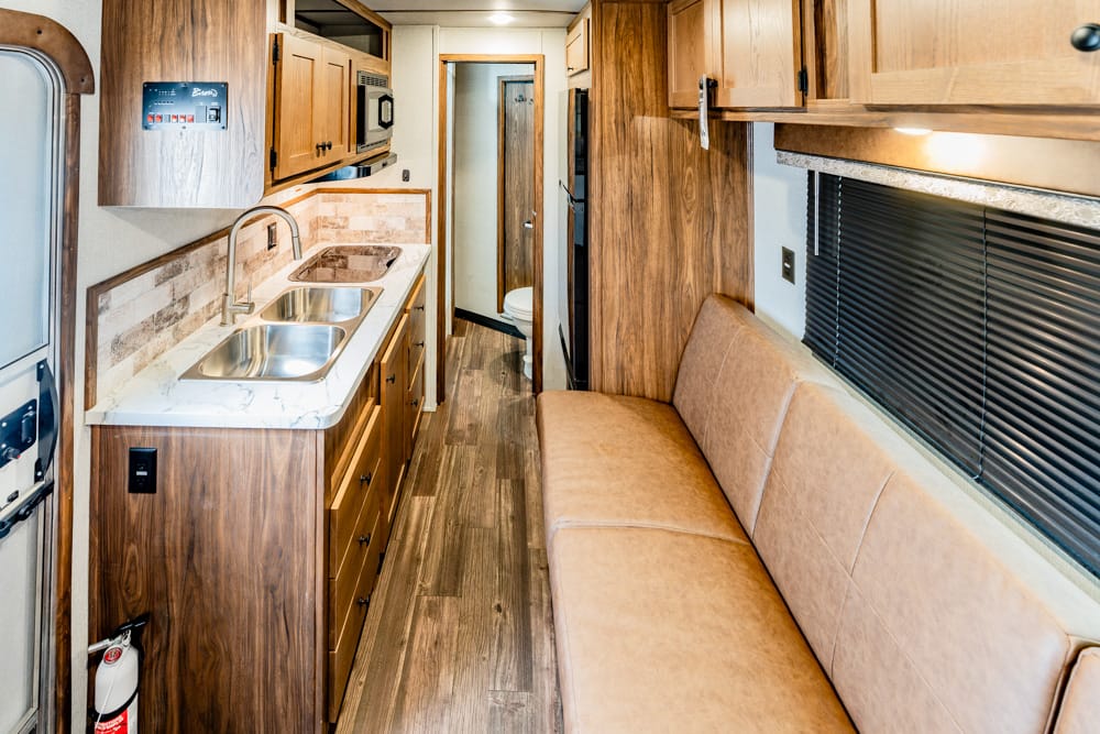 Interior of a wood-toned RV kitchen with double sink, stove, and overhead cabinets opposite a tan sofa along a window with blinds