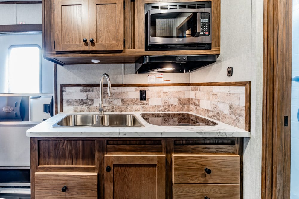 RV kitchenette with wooden cabinets, marble countertop, and a double stainless steel sink beneath a microwave
