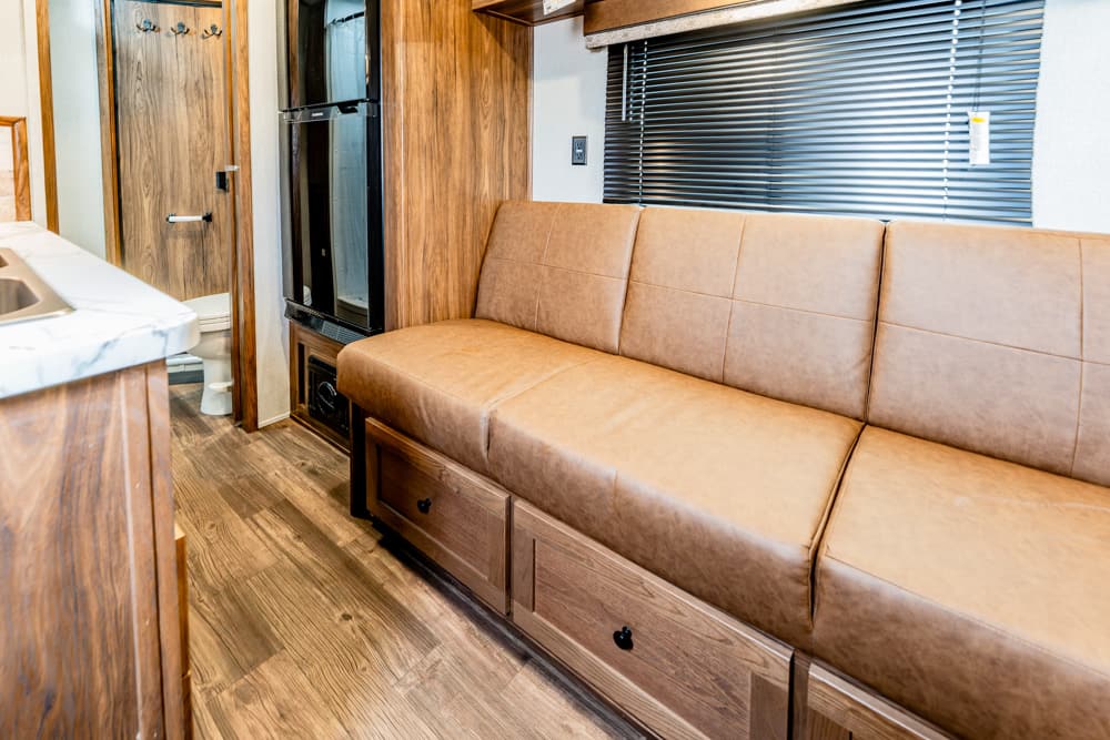RV living area with tan leather sofa and built-in storage drawers beneath, wood paneling and a window with blinds on the right side.